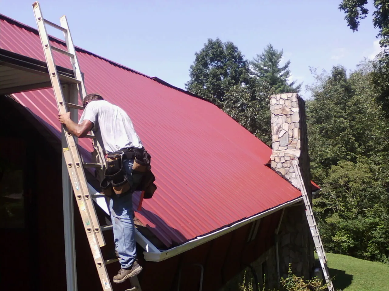 Roofer on a ladder beside a red metal roof and stone chimney — Soffit & Fascia Repair by Roofing Brothers in Kimmswick